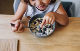 Kindergartenmädchen isst an einem Tisch ein Müsli mit Blaubeeren und zeigt auf eine Blaubeere auf dem Löffel