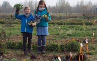 Zwei Kinder in blauen Jacken stehen auf einem Gemüsebeet und zeigen geerntete Pflanzen in die Kamera.