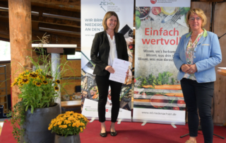 Während einer Veranstaltung stehen zwei Frauen vor Postern, wobei die linke eine Urkunde hält.
