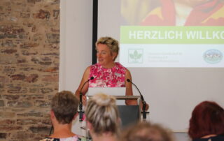 Sprecherin hinter einem Rednerpult mit zwei Mikrofonen. Im Hintergrund die Willkommensfolie der Veranstaltung.