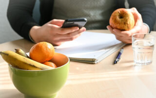 Eine Frau sitzt an einem Holztisch mit Apfel und Smartphone in der Hand. Zudem liegt ein Block mit Stift sowie ein Glas und eine Obstschale darauf.