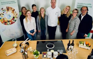 Gruppenfoto vor einer Küchenplatte mit Herd sowie verschiedenen Speisen und Getränken während einer Veranstaltung
