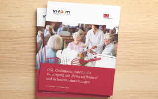 Dekoratives Foto des "DGE-Qualitätsstandards für die Gemeinschaftsverpflegung". Abgebildet auf einem Holzuntergrund.