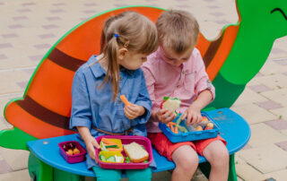 Zwei auf einer Bank sitzende Kinder essen aus Brotdosen mit Gemüse und Sandwiches