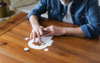 Älterer Mann sitzt am Tisch und puzzelt einen Kopf. Es fehlen noch Puzzleteile