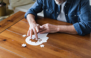 Älterer Mann sitzt am Tisch und puzzelt einen Kopf, es fehlen noch Puzzleteile