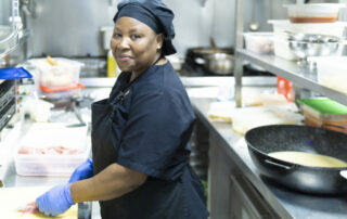 Mitarbeiterin in einer Großküche zur Kamera schauend und Fleisch schneidend